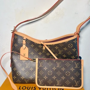 Louis Vuitton Carryall Monogram axelväska - Säljer en Louis Vuitton Carryall Monogram axelväska i klassiskt brunt och beige med ikoniskt LV-mönster. Väskan har justerbar axelrem, detaljer i ljust skinn och medföljande mindre clutch. Insidan är fodrad i vinrött tyg och har innerfack med dragkedja. Kommer med dustbag och box.