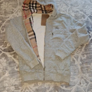 Burberry kofta - Grå hoodie från Burberry med dragkedja och huva. Insidan av huvan och öppningen har det ikoniska rutiga Burberry-mönstret i beige, svart och rött. Tröjan har broderad logga på bröstet, långa ärmar och ribbade muddar. Perfekt för en chill och stilren look.