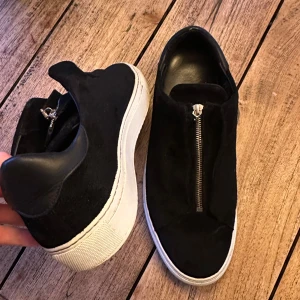 Svarta sneakers i mocka med dragkedja str 44 - Säljer ett par svarta sneakers från Arigato i mjuk mocka med vit platt sula. Skorna har en fast dragkedja. Storlek 44 men skulle rekommendera att du har något större i storlek såsom 45