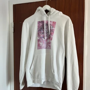 Vit hoodie från The Cool Elephant  - Vit hoodie från The Cool Elephant i bomull med en stor ficka framtill och coolt rosa och lila konsttryck på bröstet. Klassisk huva med snörning och relaxed fit. Perfekt för dig som gillar streetstyle och vill sticka ut lite extra.