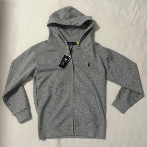 Grå hoodie från Ralph Lauren - Snygg grå hoodie från Polo Ralph Lauren med dragkedja och klassisk logga broderad på bröstet. Tröjan har huva, långa ärmar och ribbade muddar. Perfekt för chill dagar och enkel att matcha med jeans eller joggers. Skriv för mer info !