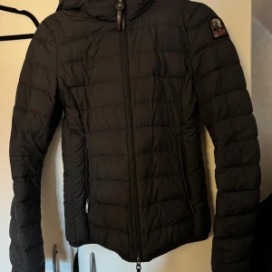 Svart dunjacka från Parajumpers - Snygg svart dunjacka från Parajumpers med huva och quiltad design. Jackan har dragkedja framtill, två sidofickor med dragkedja, den ena dragkedjan har åkt ur men går enkelt att sätta dit (kan skicka bild)och klassisk PJS-logga på ärmen. Perfekt för kyliga dagar och riktigt skön att bära. Jackan är inte svart utan mörkblå/svart. Jackan är annars i jättebra skick inga skavanker eller hål.
