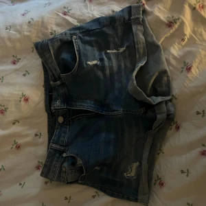 Blå jeansshorts från FB Sister - Snygga blå jeansshorts från FB Sister med slitna detaljer och klassisk femficksdesign. Shortsen har uppvikta benslut, dragkedja och knapp framtill samt läderpatch bak i midjan. Perfekta för sommaren och ger en avslappnad vibe.
