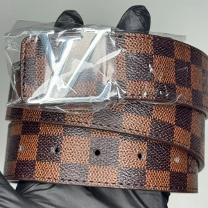Louis Vuitton rutig skinnbälte - Säljer ett snyggt LV-bälte i brunt och mörkbrunt med klassiskt rutigt mönster och en stor silverfärgad LV-spänne. Bältet är tillverkat i skinn och har flera hål för justering. Perfekt för att lyfta din outfit med en lyxig touch.