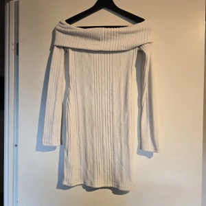 Vit ribbad offshoulder klänning - Snygg vit stickad klänning med ribbad struktur och offshoulder-design. Klänningen är kort och har långa ärmar, vilket ger en trendig och bekväm look. Perfekt för dig som gillar stilrena och enkla plagg med en twist.