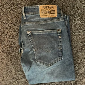 Replay blå jeans med slitningar - Säljer ett par blå jeans från Replay med snygga slitningar och lätt tvättad look. Klassisk femficksmodell med raka ben och läderpatch bak i midjan. Jeansen är i denim och har en cool, avslappnad stil som passar perfekt till sneakers.