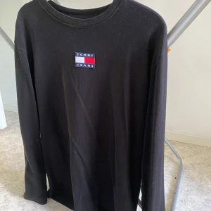 Svart långärmad tröja Tommy Jeans - Svart långärmad tröja från Tommy Jeans med klassisk rund hals och broderad logga på bröstet i rött, vitt och blått. Tillverkad i mjuk bomull som är skön mot huden. Perfekt för en clean och stilren look. Storleken är S men är stor i storlek