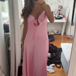 Rosa Flora Satin Maxi Dress PepperMayo - Supersnygg långklänning i satin från PepperMayo i en ljus rosa färg. Klänningen har smala axelband, v-ringning med snörning framtill och öppen rygg med rosett. Materialet är glansigt och mjukt, perfekt till bal. Modellen heter Flora Satin Maxi Dress. Orginalpris 1000kr med frakt och tull