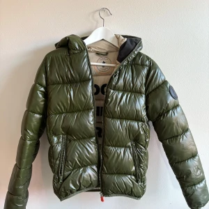 Grön pufferjacka från Save The Duck - Snygg grön pufferjacka från Save The Duck med glansig finish och huva. Jackan har dragkedja framtill, två fickor med dragkedja och svart logomärke på ärmen. Tillverkad i syntetmaterial och är helt animal free. Perfekt för kyliga dagar.