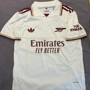 Arsenal  fotbollströja - Snygg Arsenal fotbollströja från Adidas i vit med vinröda detaljer och klassisk krage. Tröjan har korta ärmar, broderade loggor och subtila mönster i tyget. Materialet är lätt och andas, perfekt för match eller träning. Emirates och Visit Rwanda tryck på bröst och ärm.