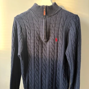 Marinblå kabelstickad tröja Polo Ralph Lauren - Kabelstickad marinblå tröja från Polo Ralph Lauren med halv dragkedja och hög krage. Tröjan har lång ärm, ribbade muddar och röd broderad logga på bröstet. Perfekt för kyliga dagar och snygg till jeans eller chinos.