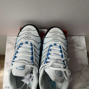 Nike Air Max Plus blå/vit sneakers - Nike Air Max Plus sneakers i vitt och blått med svarta detaljer. Skorna har vågformade linjer, synlig Air Max-dämpning och blå Swoosh på sidan. Ovandel i syntet och mesh, snörning med blå accenter och robust svart sula. Perfekta för dig som gillar streetwear.