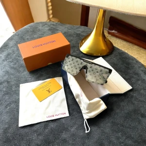 Louis Vuitton monogram solglasögon - Snygga oversized solglasögon från Louis Vuitton med svart båge och spegellins i silver. Linsen har LV:s ikoniska monogrammönster över hela ytan och detaljer i guld på skalmarna. Rektangulär, futuristisk form som verkligen sticker ut.