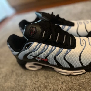 Nike Air Max Plus Tn sneakers svart/vit - Äkta Nike Air Max Plus Tn sneakers med vit mesh-överdel, svarta och blå vågformade detaljer samt svarta och röda accenter. Sulan har Tn Air-logga och röda detaljer undertill. Skorna har snörning och en sportig vibe, perfekt för dig som gillar streetwear.