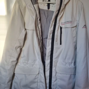 Vit vinterjacka från Everest - Säljer en snygg vit vinterjacka från Everest med huva, flera praktiska fickor och svart dragkedja. Jackan är vadderad och har meshficka på insidan. Justerbar i midjan.Perfekt för kalla dagar och har en clean look med diskreta loggor på bröst och ärm. Själva hållaren på dragkedjan trasig, men allt e helt i funktion. Se sista bilden. Använd men mycket att ge. Några små fläckar på ena fickan.