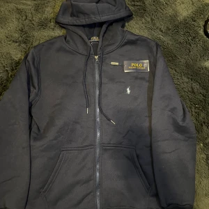 Marinblå hoodie från Polo Ralph Lauren - Snygg marinblå hoodie från Polo Ralph Lauren med dragkedja, luva och snörning. Klassisk broderad logga på bröstet och två fickor framtill. Perfekt för chill dagar eller när du vill ha en clean streetstyle-look.