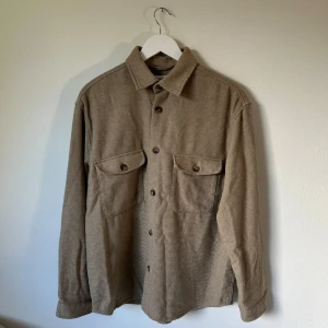 Beige overshirt från Selected Homme - Säljer en beige overshirt från Selected Homme med två stora bröstfickor och knappar framtill. Materialet är en mix av polyester och återvunnen ull som ger en mjuk och lite grov känsla. Perfekt att slänga över en t-shirt för en chill look. 3P11