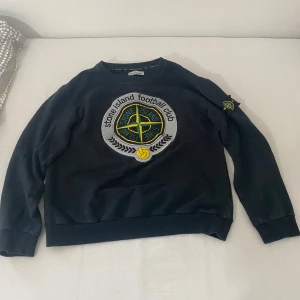 Svart Stone Island sweatshirt L - Svart sweatshirt från Stone Island med stort broderat logomärke på bröstet och klassisk patch på vänster ärm. Tröjan har rund hals, ribbade muddar och är tillverkad i mjuk bomull. Perfekt för dig som gillar streetwear och sportig stil. (Barnstorlek) 