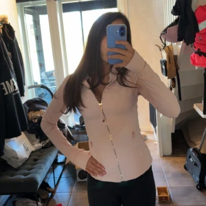 Ljusrosa ziptröja från Lululemon - Supersnygg ljusrosa ziptröja från Lululemon med figurnära passform. Tröjan har hel dragkedja framtill, två fickor med dragkedja och långa ärmar med tumhål. Perfekt för träning eller chill. Materialet är mjukt och stretchigt syntet.