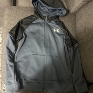 Blå och svart hoodie från Under Armour - Snygg blå hoodie från Under Armour med svarta detaljer på axlar och huva. Tröjan har dragkedja framtill, två stora fickor och en vit logga på bröstet. Perfekt för träning eller chill. Materialet är mjukt och känns sportigt.