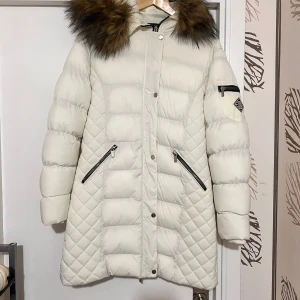 Vit lång pufferjacka Polo Club - Säljer en snygg vit pufferjacka från Polo Club med stor huva och fluffig fuskpäls. Jackan har quiltade partier, dragkedjor på fickorna och ärmarna samt en badge på ena ärmen. Perfekt för kalla dagar och riktigt mysig att bära.