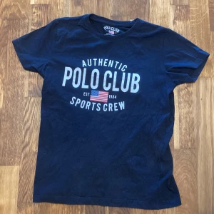 Mörkblå Polo Club t-shirt - Snygg mörkblå t-shirt från Polo Club med stort tryck framtill och amerikansk flagga. Klassisk rund hals och korta ärmar. Perfekt för dig som gillar sportig och avslappnad stil. Tillverkad i mjuk bomull för skön känsla hela dagen.