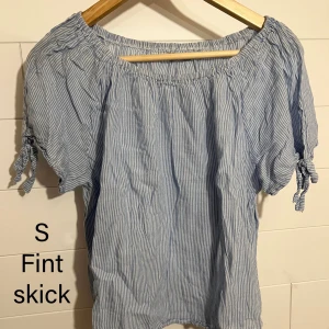 Blåvit randig blus med knytärmar - Söt blus i blåvitt randigt med korta ärmar och knyt vid ärmsluten. Blusen har en avslappnad passform och resår vid halsringningen för en luftig känsla. Perfekt för varma dagar och enkel att styla till jeans eller kjol.