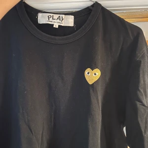 Svart Comme des Garçons Play t-shirt - Svart t-shirt från Comme des Garçons Play med klassisk rund hals och korta ärmar. På bröstet sitter det ikoniska hjärtat i gult/guld med ögon. T-shirten är i mjuk bomull och har en clean, stilren look.