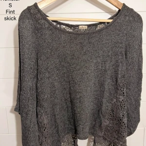 Grå stickad tröja från Hollister - Grå stickad tröja från Hollister med vida ärmar och snygga virkade spetsdetaljer med blommönster längs nederkant och rygg. Rund halsringning och mjukt material.