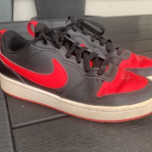 Nike sneakers svart/röd strl 36.5 - Säljer ett par feta Nike sneakers i svart och rött med klassisk swoosh på sidan. Skorna har snörning, rund tå och platt sula. Ovandelen är i skinn och textil, sulan är röd och vit. Perfekta för dig som gillar streetstyle och vill sticka ut.