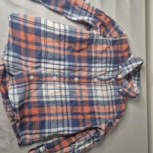 Rutig skjorta från Lindex i bomull - Säljer en rutig skjorta från Lindex i mjuk bomull. Skjortan har blå, orange och vita rutor, klassisk krage och knäppning framtill. Perfekt för dig som gillar en avslappnad och stilren look. Långärmad modell med bekväm passform.