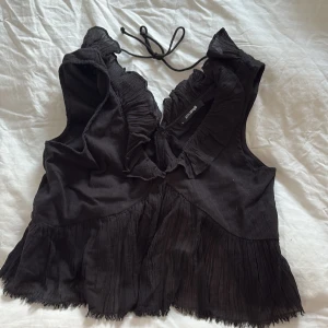 Svart volangblus med knyt från Bik Bok - Supersöt svart blus med volangdetaljer och knytning i nacken och ryggen. Blusen är ärmlös och har en v-ringad front med rynkade volanger längs kanterna. 
