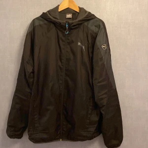 Svart vindjacka från Puma XL - Svart vindjacka från Puma i storlek XL med huva och dragkedja. Jackan har meshfoder, logga på bröstet och ärmen samt detaljer i grått. Perfekt för blåsiga dagar och sportiga outfits.