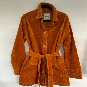 Manchesterjacka med bälte från Monki - Säljer en snygg orange manchesterjacka från Monki i storlek XXS. Jackan har bälte i midjan, stora fickor framtill och knäpps med knappar. Perfekt för dig som gillar retrovibbar och vill sticka ut lite extra.🤩💕