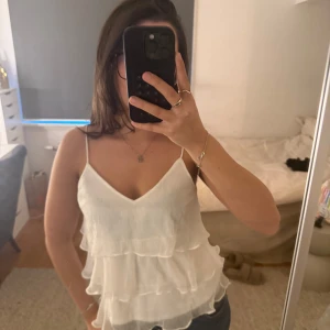 Vit volangig linnetopp med tunna axelband - Säljer en superfin vit linnetopp med flera lager volanger och tunna axelband. Toppen har v-ringning och är perfekt för varma dagar. Den är lätt och luftig, vilket gör den riktigt skön att bära till jeans eller kjol.