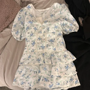 Blommig volangklänning Design by Amor - Supersöt kort klänning från Design by Amor med blått blommönster på vit botten. Puffärmar, spetsdetaljer och flera volanger. Perfekt för sommarens alla tillfällen. V-ringad och med fina detaljer framtill.