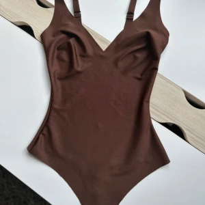 Brun v-ringad bodysuit av skims med justerbara band - Snygg brun bodysuit av märket skims i hel modell med v-ringad hals och justerbara axelband. Materialet är mjukt och stretchigt, troligen syntet. Praktisk knäppning i grenen för enkel användning. Perfekt för dig som vill ha en stilren look