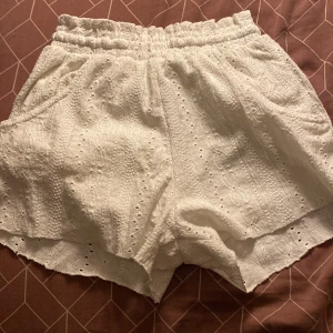 Vita broderade shorts från Zara - Superfina vita shorts från Zara med broderade detaljer och elastisk midja. De har en luftig passform, fickor på sidorna och är tillverkade i ett mjukt bomullstyg. Perfekta för varma sommardagar och ger en söt och trendig look.