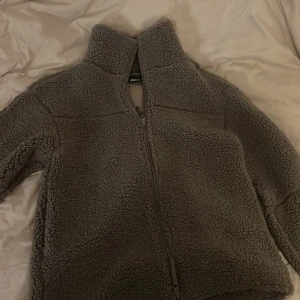 Ljus brun teddyjacka från Gina Tricot - Mysig brun teddyjacka från Gina Tricot med hög krage och hel dragkedja framtill. Jackan är tillverkad i fluffigt fleecematerial och har långärmad passform. Perfekt för kalla dagar när du vill vara både varm och snygg.