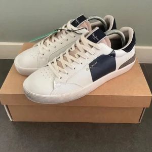 Pepe Jeans vita och marinblå sneakers - Snygga sneakers från Pepe Jeans i vitt med marinblå och beige detaljer. Skorna har snörning, rund tå och platt sula. Ovandelen är i skinn och mocka, med logga på sidan och '1973' präglat på hälen. Perfekta för en stilren och avslappnad look.