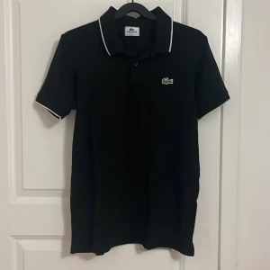 Svart pikétröja Lacoste - Svart pikétröja Lacoste med klassisk krage och vita detaljer på krage och ärmslut. Ikoniska gröna krokodilloggan på bröstet. Skön passform och tillverkad i bomull, perfekt för en clean och sportig look.