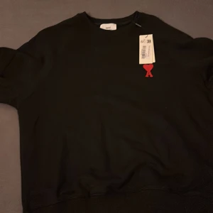 Svart sweatshirt från Ami Paris - Svart sweatshirt från Ami Paris med klassisk rund hals och röd broderad Ami de Coeur-logo på bröstet. Tröjan är i mjuk bomull och har en clean, stilren look som passar perfekt till jeans eller dressade byxor.