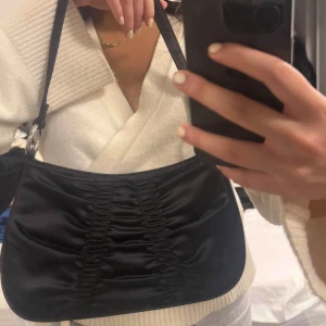 Svart axelväska i satin med rynk - Snygg svart axelväska i glansigt satinmaterial med rynkade detaljer på framsidan. Väskan har justerbar axelrem och silvriga ringdetaljer. Perfekt storlek för det viktigaste och ger en trendig vibe till din outfit.