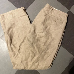 Beiga linnebyxor från Joyboy - Snygga beiga linnebyxor från Joyboy, tillverkade i 100% linne. Byxorna har klassisk passform med hällor för bälte, knapp och dragkedja i midjan samt bakficka med knapp. Fläck längst ned på byxan som säkert går bort i tvätten