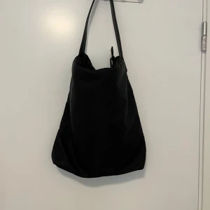 Svart axelväska i syntetmaterial - Stilren svart axelväska med enkel design och långa handtag. Väskan är tillverkad i mjukt syntetmaterial och har en rymlig form, perfekt för att bära med sig allt man behöver. Stängs med dragkedja upptill och passar till många olika outfits.