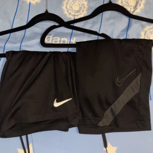 Svarta Nike träningsshorts - Två par svarta träningsshorts från Nike med klassisk logga på framsidan. Ena paret har en diskret grå detalj längs sidan. Perfekta för gymmet eller löprundan, med bekväm passform och lätt material som andas.
