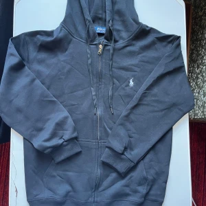 Svart hoodie från Polo Ralph Lauren - Svart hoodie från Polo Ralph Lauren med dragkedja och huva. Klassisk design med broderad logga på bröstet, ribbade muddar och fickor framtill. Tillverkad i 100% bomull för en skön och avslappnad känsla.