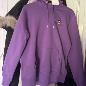 Lila hoodie från GANT med logga - Säljer en lila hoodie från GANT med broderad logga på bröstet. Tröjan har huva med snörning och känguruficka. Helt oanvänd och perfekt för kyliga dagar. 