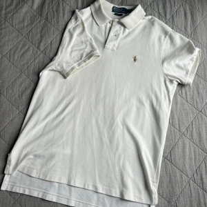 Cremevit polo t-shirt från Ralph Lauren - Cremevit polo t-shirt från Ralph Lauren i klassisk custom fit. T-shirten har korta ärmar, krage med två knappar och den  broderade loggan på bröstet i guld. Tillverkad i mjuk bomull. Väldigt schysst pikétröja!