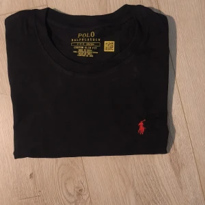Svart t-shirt Polo Ralph Lauren S - Snygg svart t-shirt från Polo Ralph Lauren med röd broderad logga på bröstet. Modellen är slim fit och har klassisk rund hals. Tillverkad i 100% mjuk bomull, perfekt för en clean och stilren look. Storlek S. Mått: 44 cm över bröstet, 67 cm längd. Helt oanvänd i nytt skick✨